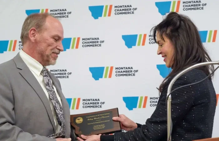 Secretary Christi Jacobsen recognizes 100-year businesses during Montana Chamber of Commerce Business Days at the Capitol 9 A man in a suit receives a plaque from a woman during an event, with a Montana Chamber of Commerce backdrop behind them. Both are smiling and making eye contact.