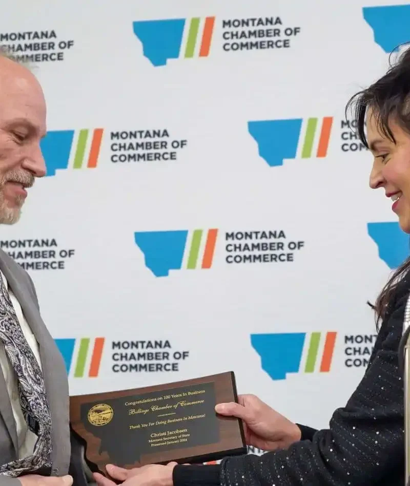 A man in a suit receives a plaque from a woman during an event, with a Montana Chamber of Commerce backdrop behind them. Both are smiling and making eye contact.
