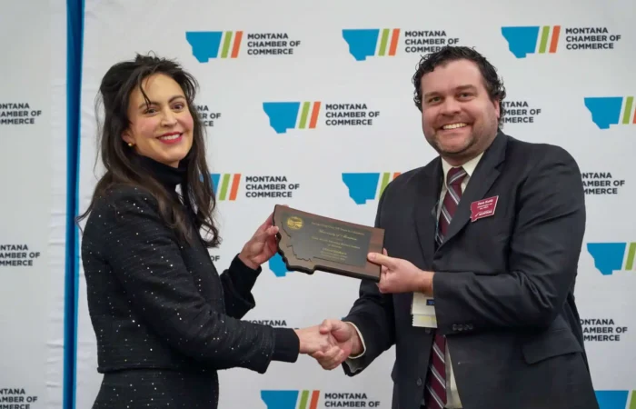 Secretary Christi Jacobsen standing up for small businesses, monitoring Corporate Transparency Act court decision 7 Two people in business attire shake hands and smile as one presents a plaque to the other, celebrating small businesses. They stand in front of a Montana Chamber of Commerce backdrop.