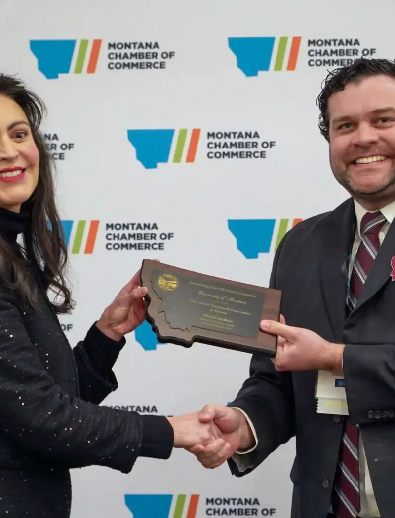 Two people in business attire shake hands and smile as one presents a plaque to the other, celebrating small businesses. They stand in front of a Montana Chamber of Commerce backdrop.
