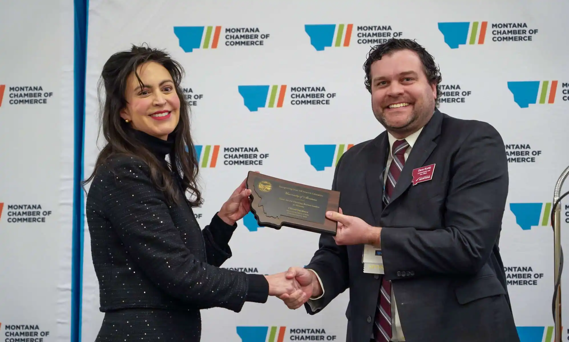 Two people in business attire shake hands and smile as one presents a plaque to the other, celebrating small businesses. They stand in front of a Montana Chamber of Commerce backdrop.