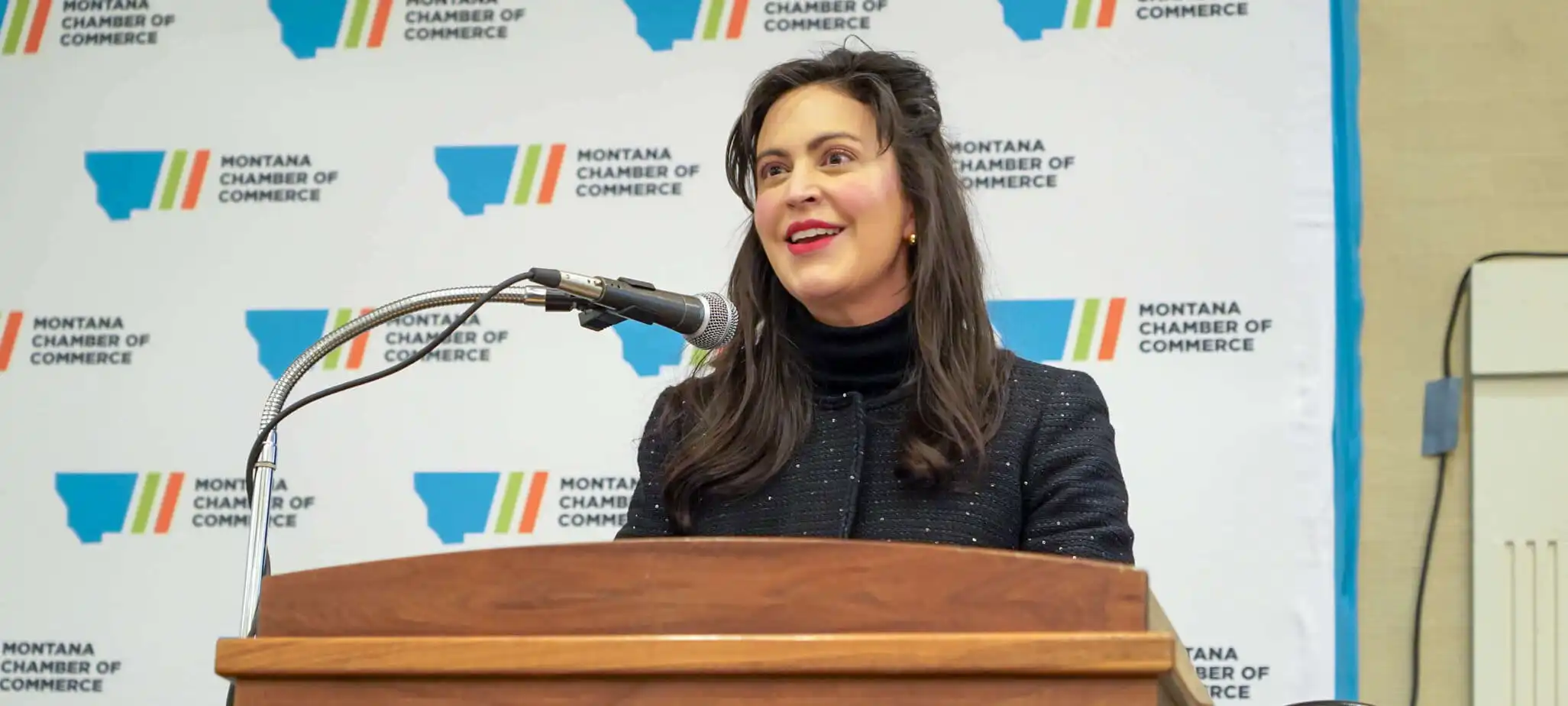 Christi Jacobsen, with long dark hair, smiles as she speaks at a podium in front of a backdrop featuring the Montana Chamber of Commerce logo during Business Days.