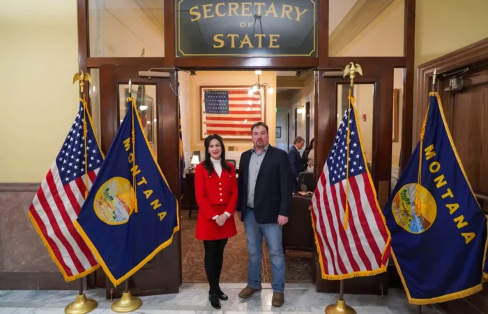Secretary Christi Jacobsen: 'Nearly 200 candidate filings processed to date' 1 Two people stand side by side in front of the Montana Secretary of State office entrance, with U.S. and Montana flags on either side and a large American flag in the background.