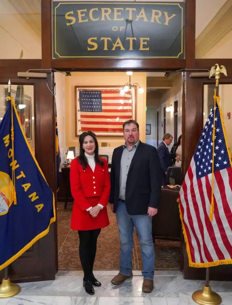 Two people stand side by side in front of the Montana Secretary of State office entrance, with U.S. and Montana flags on either side and a large American flag in the background.