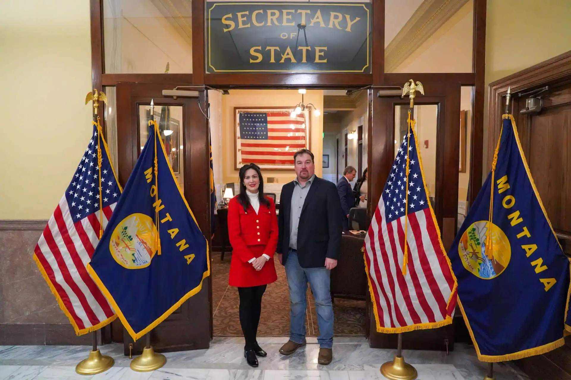 Two people stand side by side in front of the Montana Secretary of State office entrance, with U.S. and Montana flags on either side and a large American flag in the background.