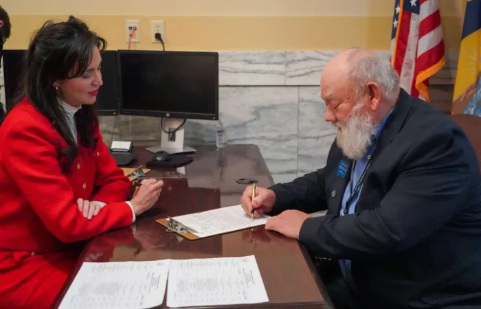 2024 Candidate Filing photo gallery 3 A woman in a red suit and a man with a white beard in a dark suit sit at a desk; the man is signing a document while the woman watches. Two monitors and American flags are visible in the background.