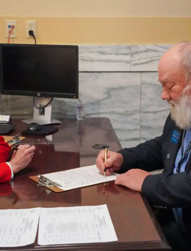 A woman in a red suit and a man with a white beard in a dark suit sit at a desk; the man is signing a document while the woman watches. Two monitors and American flags are visible in the background.