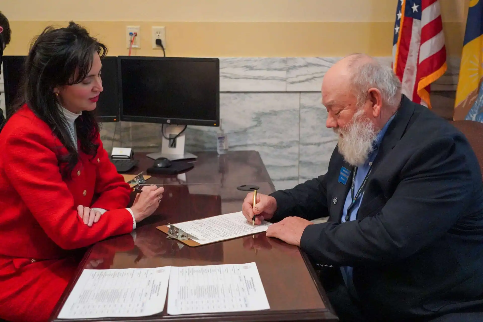 A woman in a red suit and a man with a white beard in a dark suit sit at a desk; the man is signing a document while the woman watches. Two monitors and American flags are visible in the background.