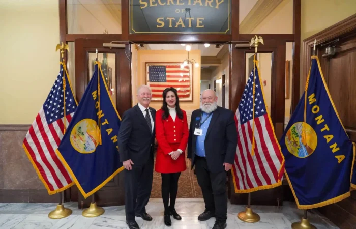 Secretary Christi Jacobsen: 'More than 100 candidates file on opening day of 2024 candidate filing' 2 Three people stand smiling in front of a Secretary of State sign, flanked by U.S. and Montana flags. The woman in the center wears a red outfit; the two men on either side wear suits. An American flag hangs in the background.