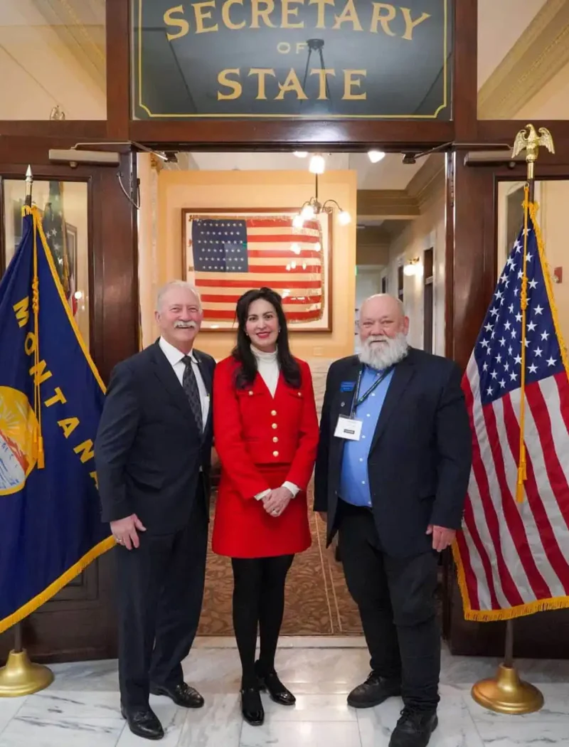 Three people stand smiling in front of a Secretary of State sign, flanked by U.S. and Montana flags. The woman in the center wears a red outfit; the two men on either side wear suits. An American flag hangs in the background.
