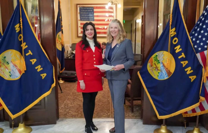 Secretary Christi Jacobsen, Secretary of State's Office open 2024 candidate filing 4 Two women stand smiling between two Montana state flags in a formal room with an American flag in the background. Secretary of State Christi Jacobsen, in a gray suit holding papers, joins a guest as they mark the 2024 candidate filing event.