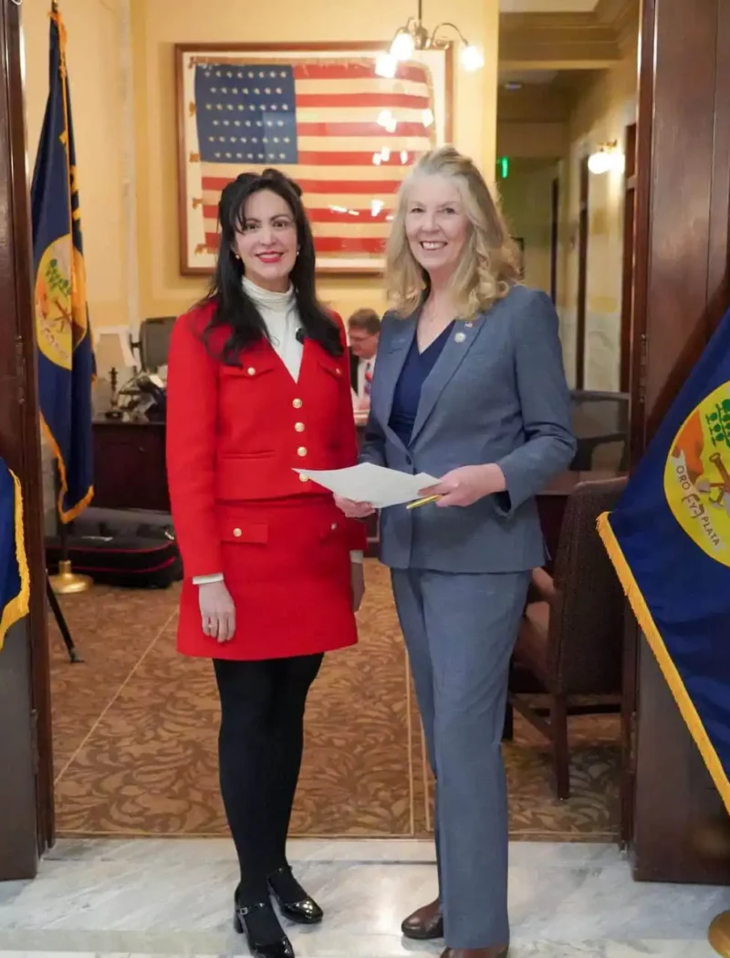 Two women stand smiling between two Montana state flags in a formal room with an American flag in the background. Secretary of State Christi Jacobsen, in a gray suit holding papers, joins a guest as they mark the 2024 candidate filing event.