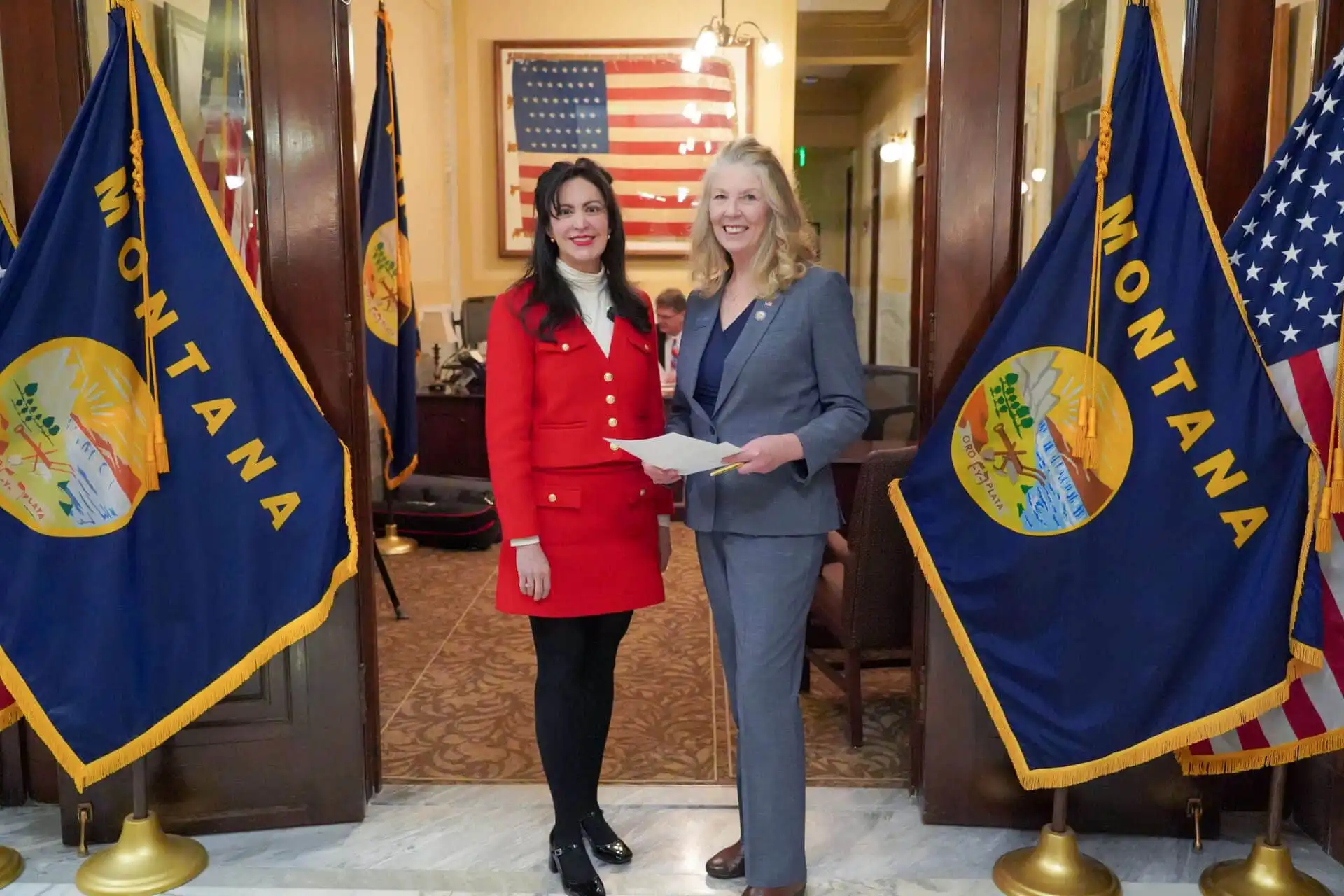 Two women stand smiling between two Montana state flags in a formal room with an American flag in the background. Secretary of State Christi Jacobsen, in a gray suit holding papers, joins a guest as they mark the 2024 candidate filing event.