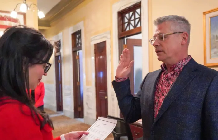 Secretary Christi Jacobsen administers swearing-in of new Montana Director of Commerce Paul Green 8 A man wearing glasses and a patterned shirt raises his right hand to take an oath as a woman in red holds a document, inside a formal hallway with marble walls and wooden doors.