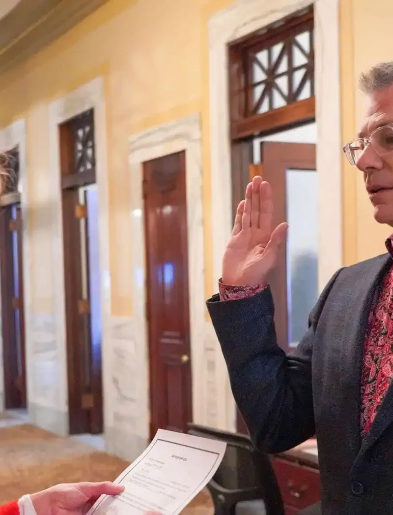 A man wearing glasses and a patterned shirt raises his right hand to take an oath as a woman in red holds a document, inside a formal hallway with marble walls and wooden doors.