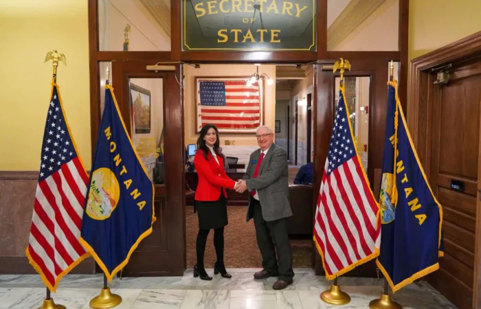 Secretary Jacobsen officially opens Montana's 2022 candidate filing 4 Two people shake hands in front of a “Secretary of State” sign, flanked by two U.S. flags and two Montana state flags, as Secretary Jacobsen oversees the 2022 candidate filing inside her Montana office. An American flag hangs in the background.