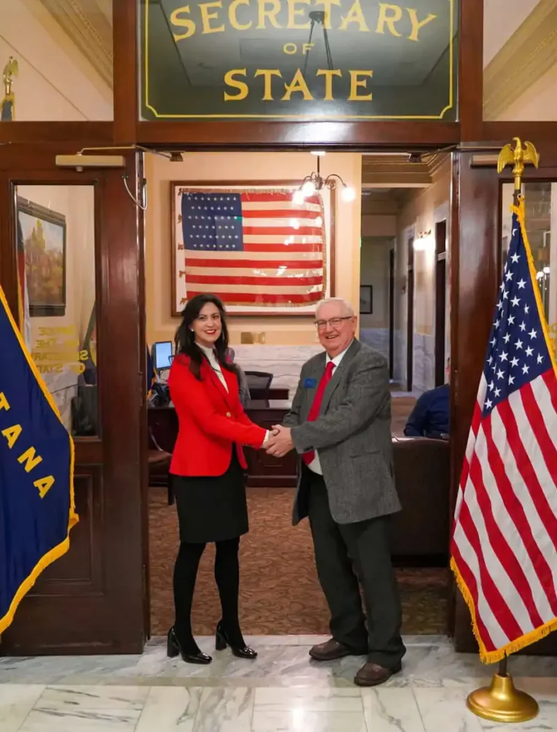 Two people shake hands in front of a “Secretary of State” sign, flanked by two U.S. flags and two Montana state flags, as Secretary Jacobsen oversees the 2022 candidate filing inside her Montana office. An American flag hangs in the background.