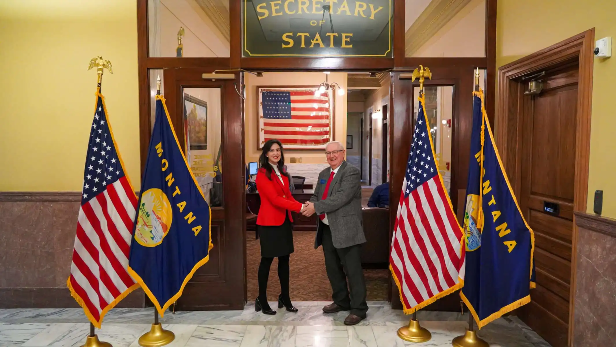 Secretary Jacobsen officially opens Montana's 2022 candidate filing 8 Two people shake hands in front of a “Secretary of State” sign, flanked by two U.S. flags and two Montana state flags, as Secretary Jacobsen oversees the 2022 candidate filing inside her Montana office. An American flag hangs in the background.