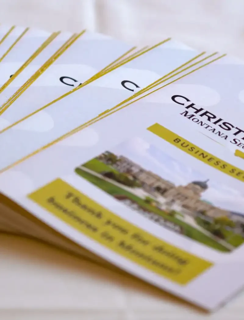 A stack of brochures titled “Christi Jacobsen Montana Secretary of State Business Services,” featuring a seal and a photo of the Montana State Capitol on the front, arranged neatly on a white tablecloth.