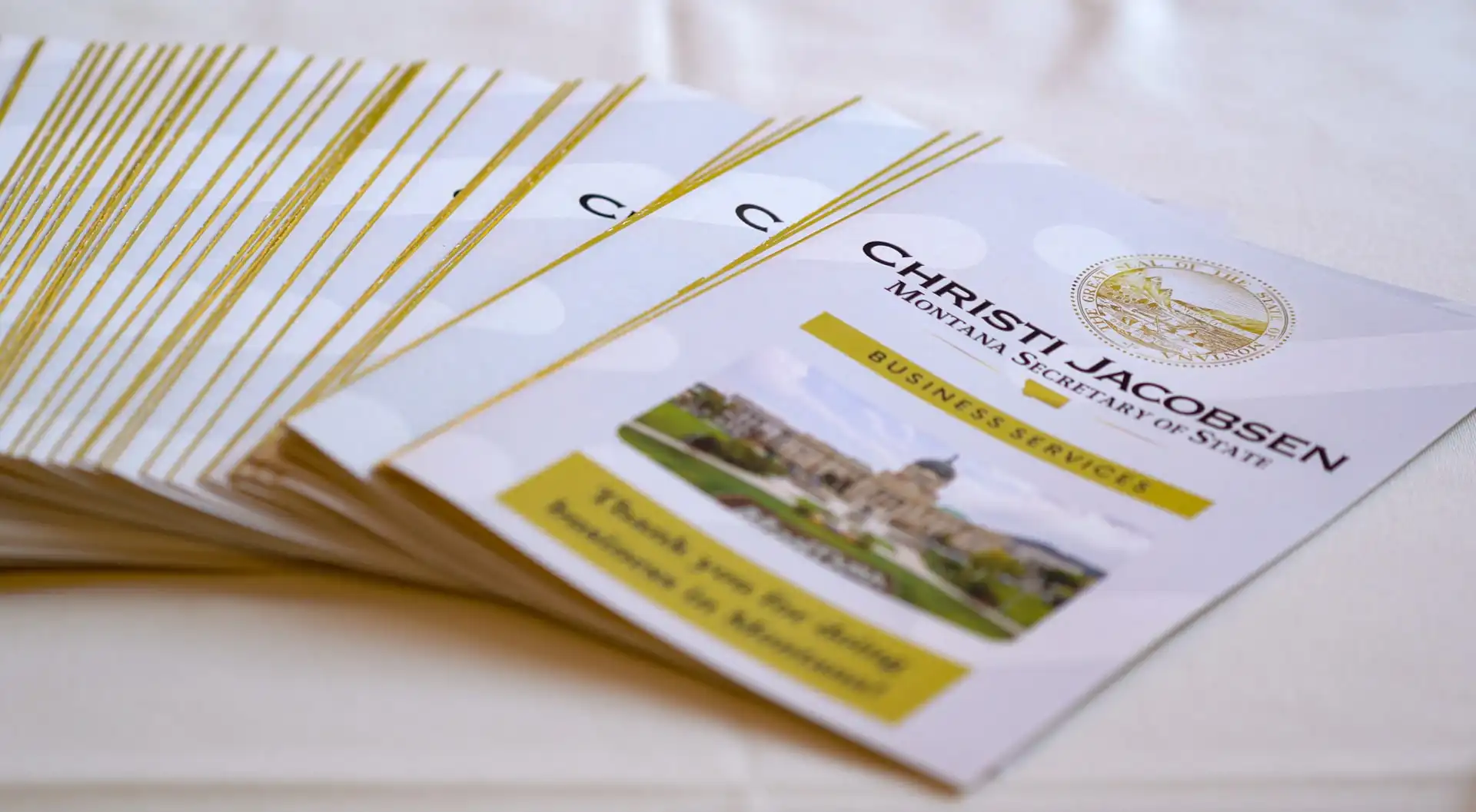 A stack of brochures titled “Christi Jacobsen Montana Secretary of State Business Services,” featuring a seal and a photo of the Montana State Capitol on the front, arranged neatly on a white tablecloth.