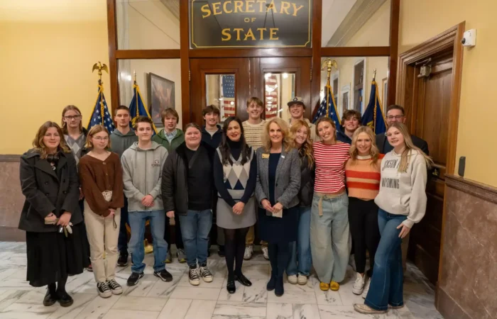 Stevensville students visit Montana State Capitol, Secretary of State's Office 10 A group of young adults and two women in business attire, including Stevensville students, pose together in front of a door labeled Secretary of State, with American flags and marble floors visible at the Montana State Capitol.