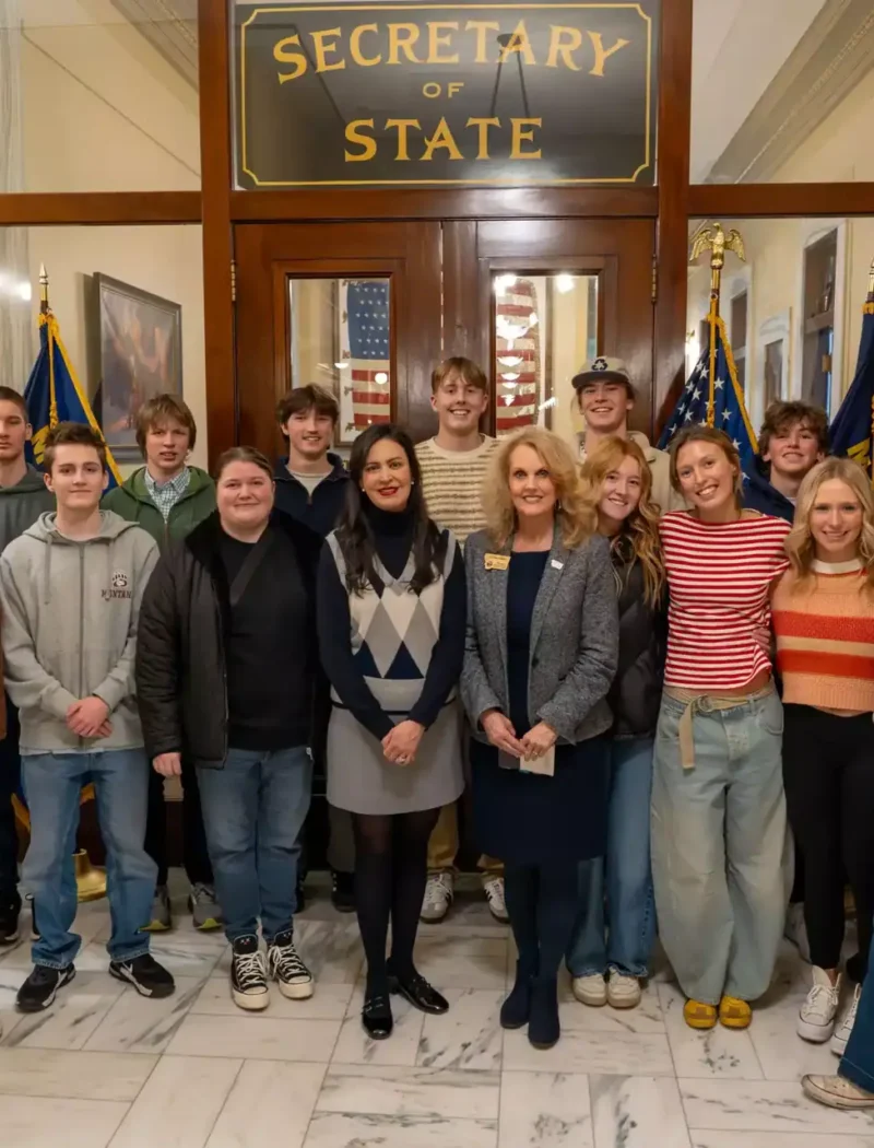 A group of young adults and two women in business attire, including Stevensville students, pose together in front of a door labeled Secretary of State, with American flags and marble floors visible at the Montana State Capitol.