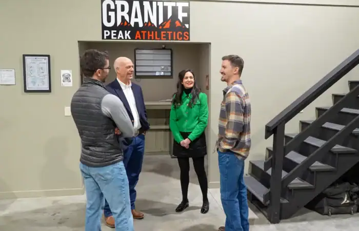 Four people stand in conversation near a staircase inside Granite Peak Athletics, a Montana business. Two men face away while one man and one woman, who may be local athletes, smile at the camera under the sign.