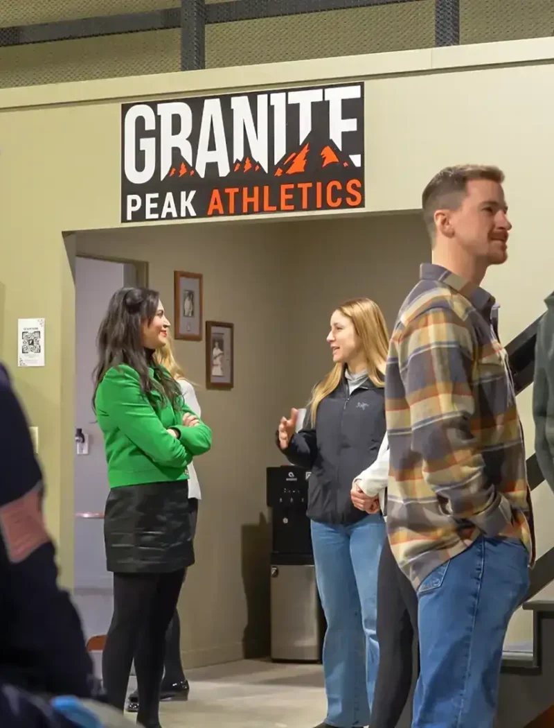 A group of people stands and talks inside a building near a Granite Peak Athletics sign. Two women stand near the doorway while others, including people in blue uniforms, converse nearby during National Small Business Month.
