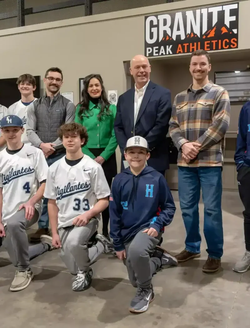 A youth baseball team and five adults pose indoors in front of a Granite Peak Athletics sign. The players wear Highlanders uniforms and caps, while the adults stand behind them, smiling—celebrating community and business creation in 2024.