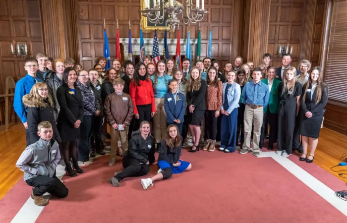 Secretary Christi Jacobsen visits with Boy Scout, 4-H members 4 A large group of young people and adults, including 4-H members and Boy Scouts, pose together in a wood-paneled room with flags, a chandelier, and large windows. Most are dressed in formal or semi-formal clothing and are smiling at the camera.