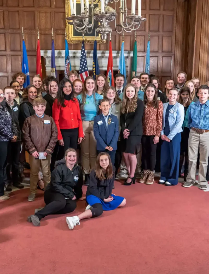 A large group of young people and adults, including 4-H members and Boy Scouts, pose together in a wood-paneled room with flags, a chandelier, and large windows. Most are dressed in formal or semi-formal clothing and are smiling at the camera.