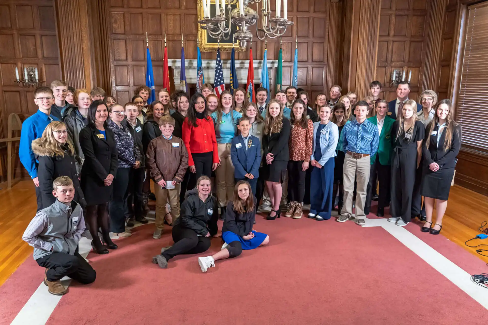 A large group of young people and adults, including 4-H members and Boy Scouts, pose together in a wood-paneled room with flags, a chandelier, and large windows. Most are dressed in formal or semi-formal clothing and are smiling at the camera.