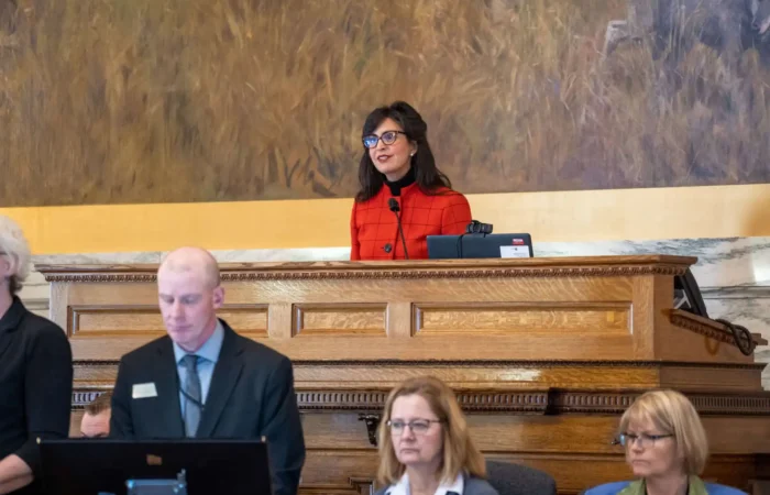 Secretary Christi Jacobsen addresses Montana House of Representatives to open 68th Legislative Session 6 A woman in a red top stands at a podium, speaking and smiling during the 68th Legislative Session, while three seated people, two women and a man, focus on documents or screens in front of her in the Montana House of Representatives.