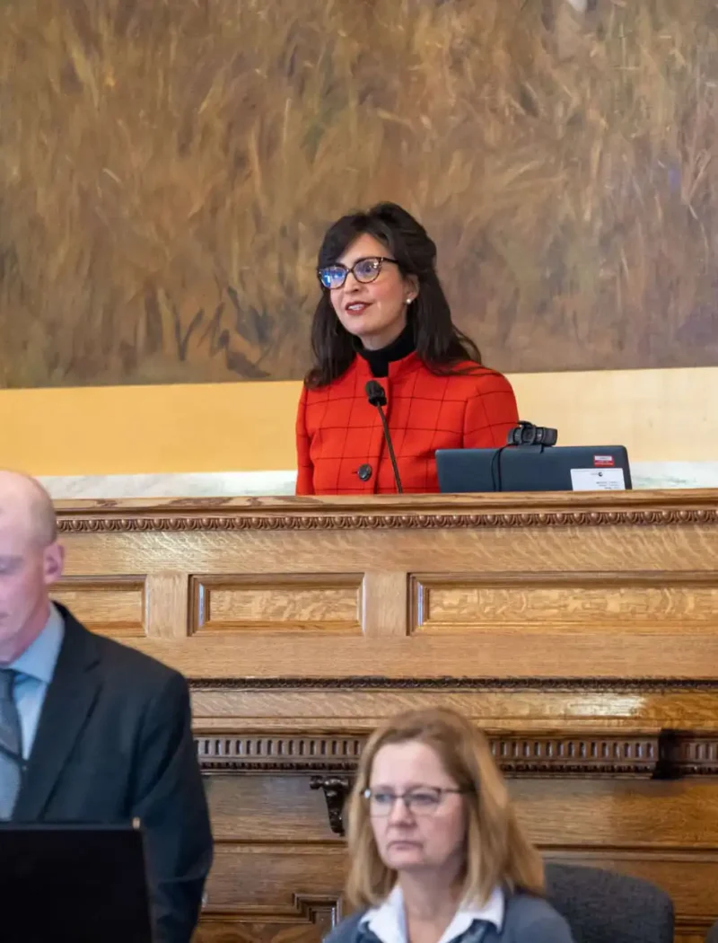 A woman in a red top stands at a podium, speaking and smiling during the 68th Legislative Session, while three seated people, two women and a man, focus on documents or screens in front of her in the Montana House of Representatives.