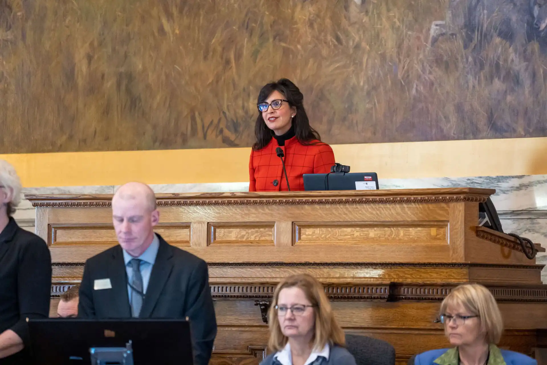 A woman in a red top stands at a podium, speaking and smiling during the 68th Legislative Session, while three seated people, two women and a man, focus on documents or screens in front of her in the Montana House of Representatives.