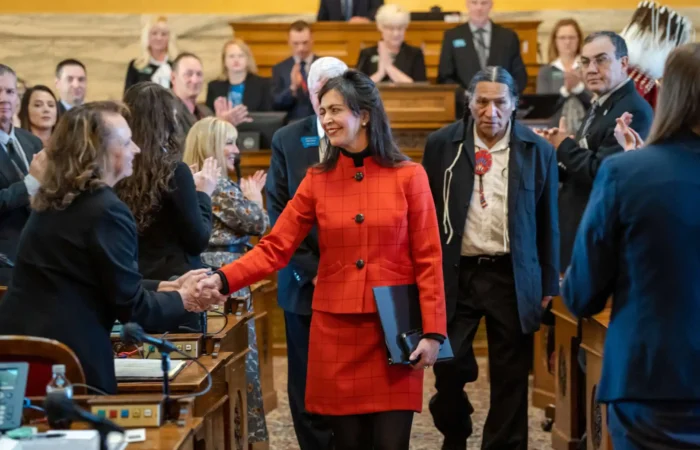 During the opening week of the Montana Legislative Session, a woman in a red suit smiles and shakes hands with another person in a formal assembly room as people stand, applaud, and photos are taken. She holds a folder while others look on.