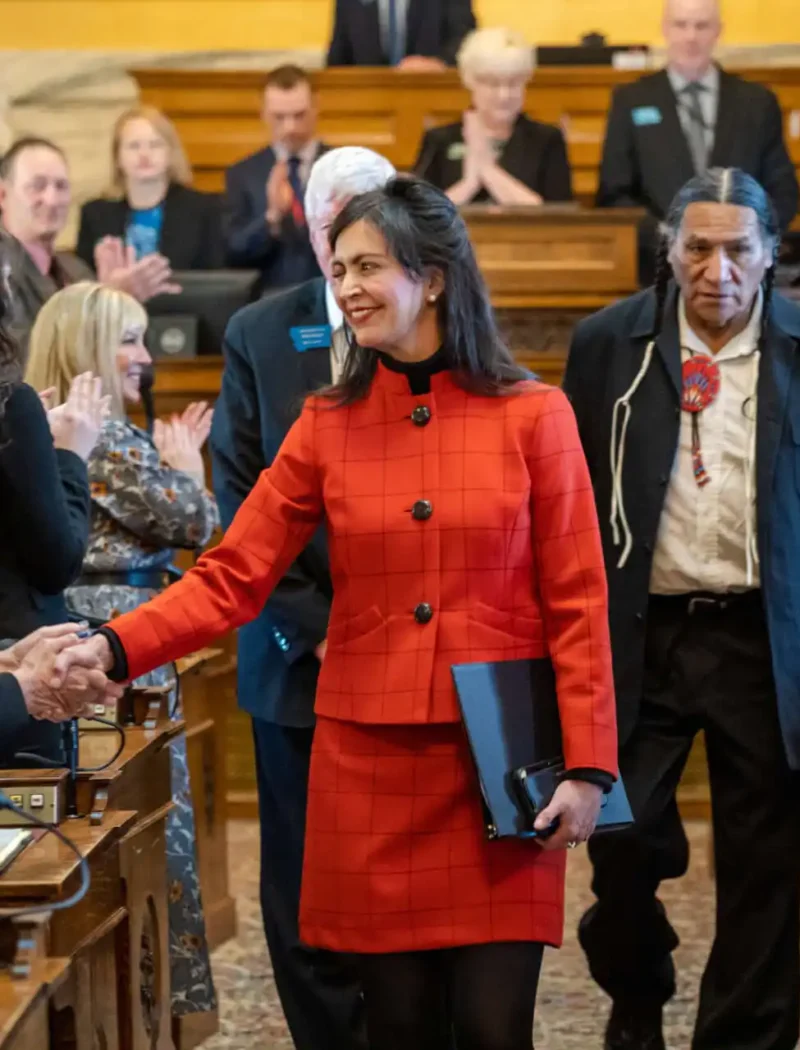 During the opening week of the Montana Legislative Session, a woman in a red suit smiles and shakes hands with another person in a formal assembly room as people stand, applaud, and photos are taken. She holds a folder while others look on.