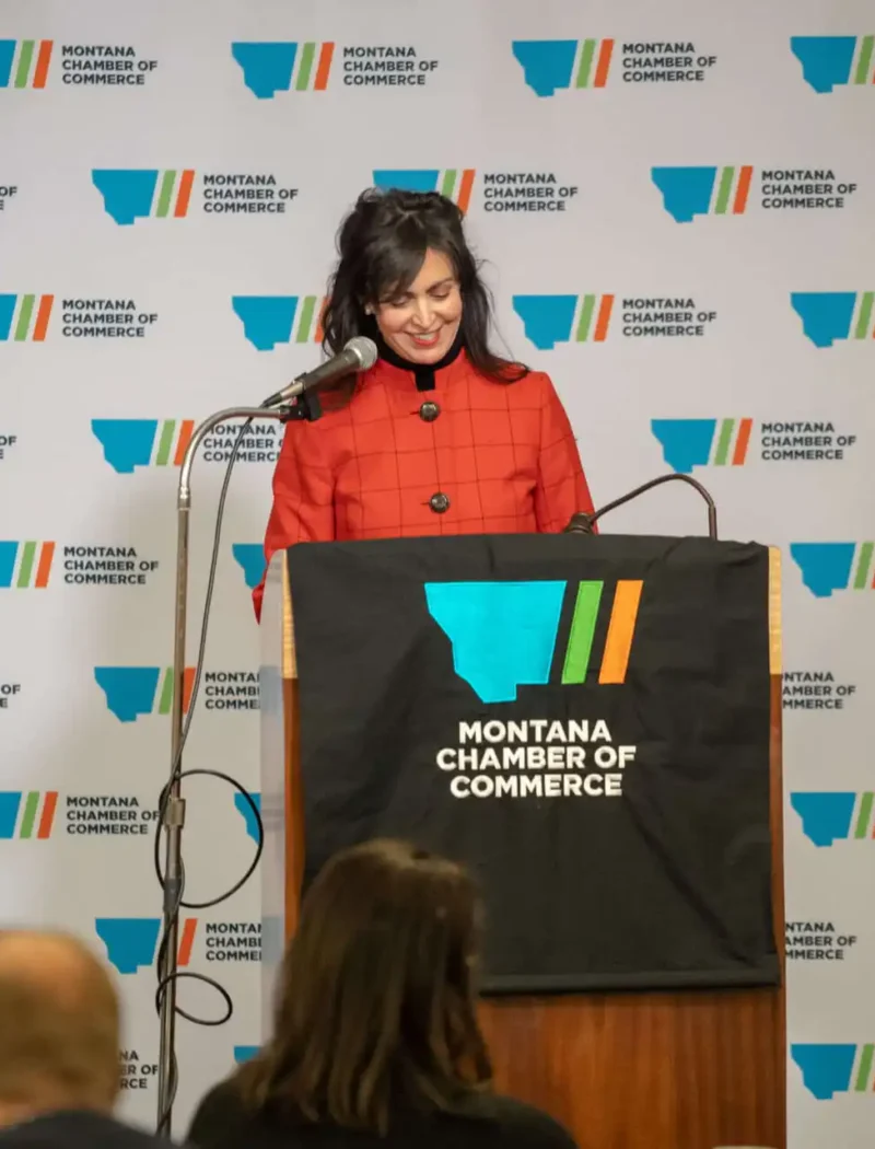 Secretary Christi Jacobsen recognizes Montana local businesses during National Small Business Week 1 A woman in a red jacket stands at a podium with a Montana Chamber of Commerce sign, speaking during the Montana Legislative Session opening week. A branded backdrop and several attendees are visible in these event photos.