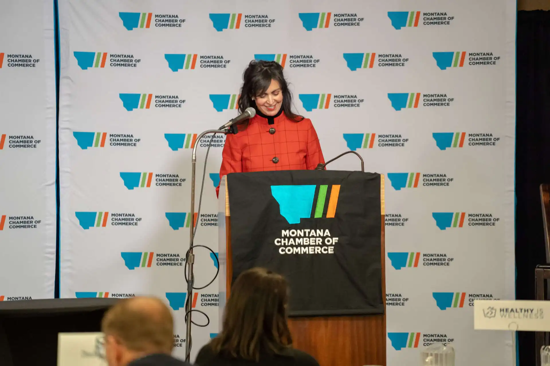 A woman in a red jacket stands at a podium with a Montana Chamber of Commerce sign, speaking during the Montana Legislative Session opening week. A branded backdrop and several attendees are visible in these event photos.
