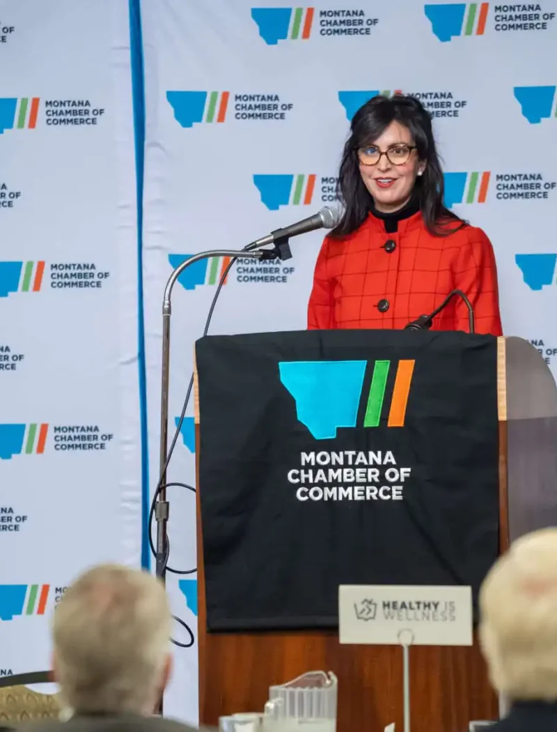 A woman in a red jacket speaks at a podium covered with a “Montana Chamber of Commerce” banner, addressing an audience at an indoor event. The background has a repeating Chamber of Commerce logo.