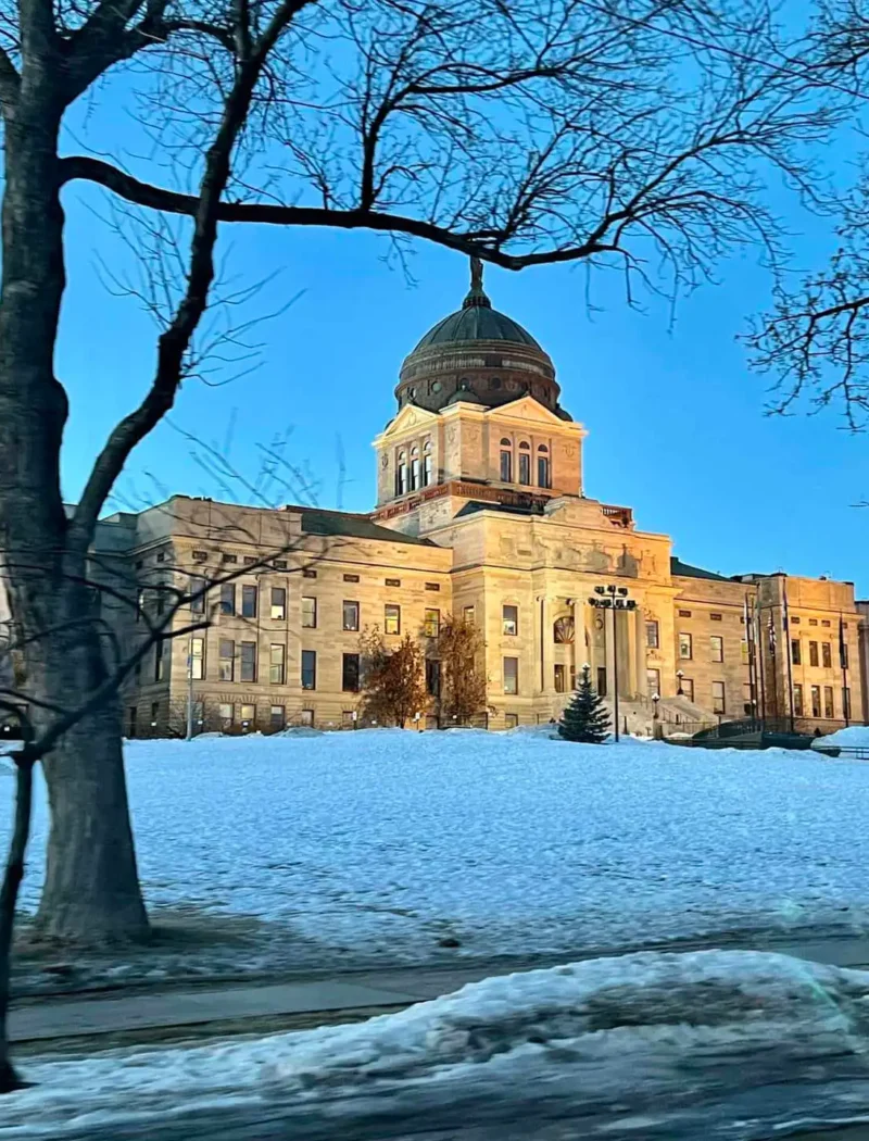 A large domed government building is illuminated at dusk, surrounded by snow-covered ground and bare trees, with a clear blue sky in the background.