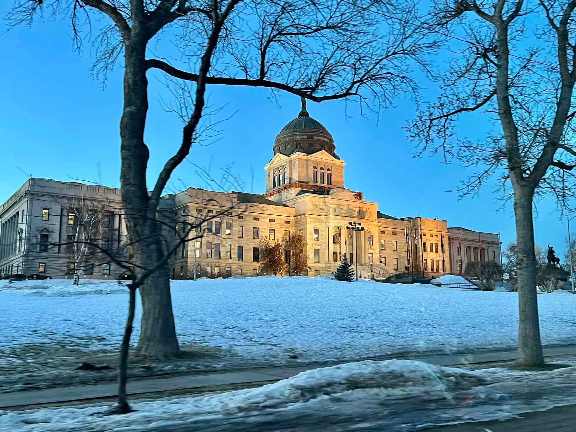 A large domed government building is illuminated at dusk, surrounded by snow-covered ground and bare trees, with a clear blue sky in the background.