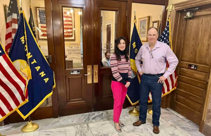 Secretary Christi Jacobsen: '220 candidate filings processed as of January 26' 10 Two people stand smiling in front of wooden doors at a government building, flanked by Montana and U.S. flags. The woman wears bright pink pants and a striped sweater, and the man wears a light pink shirt and blue jeans.