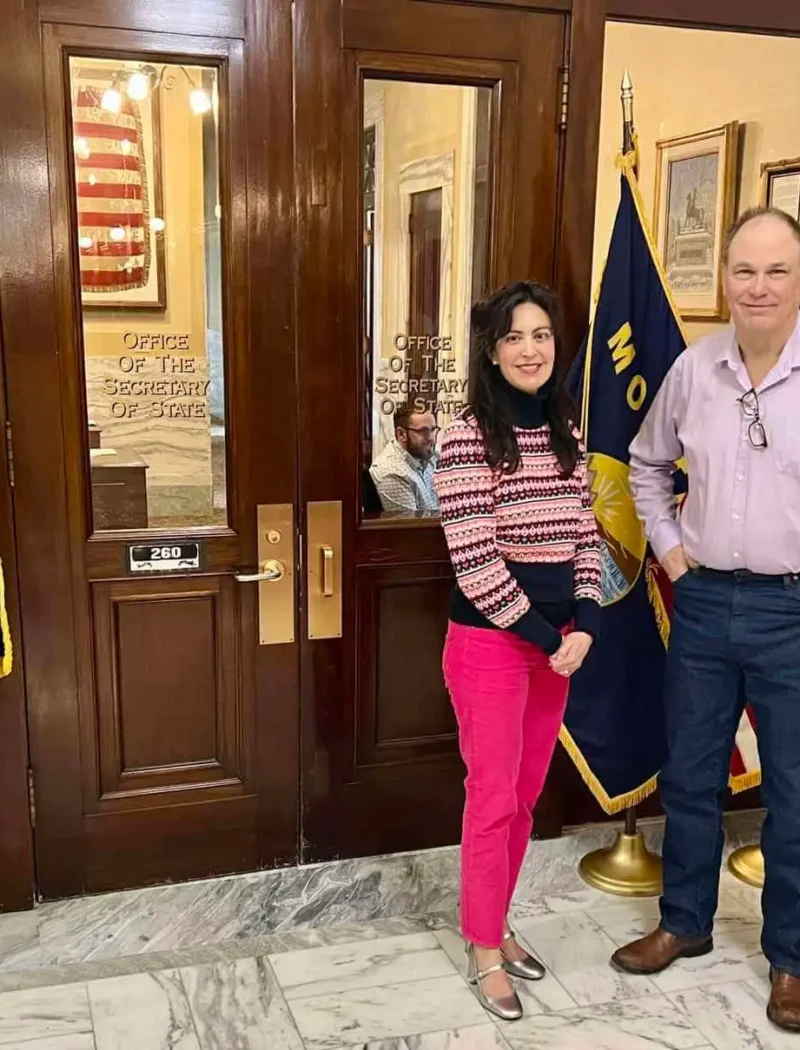 Two people stand smiling in front of wooden doors at a government building, flanked by Montana and U.S. flags. The woman wears bright pink pants and a striped sweater, and the man wears a light pink shirt and blue jeans.