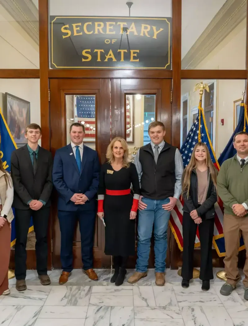 A group of seven Lima students stand in a line, posing for a photo in front of the Montana Secretary of State office entrance, flanked by U.S. and state flags on both sides.
