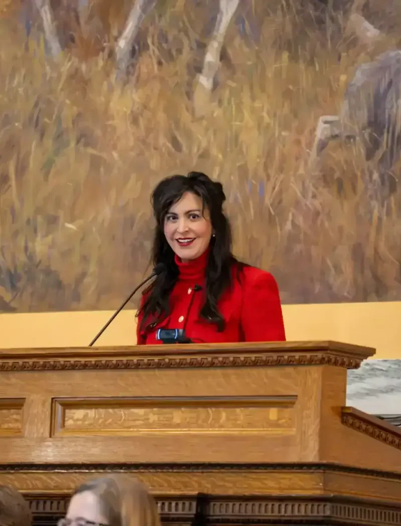 Secretary of State Christi Jacobsen, wearing a red blouse, stands at a wooden podium with microphones, smiling in front of a mural featuring animals and tall grass during the 69th Legislative Assembly.