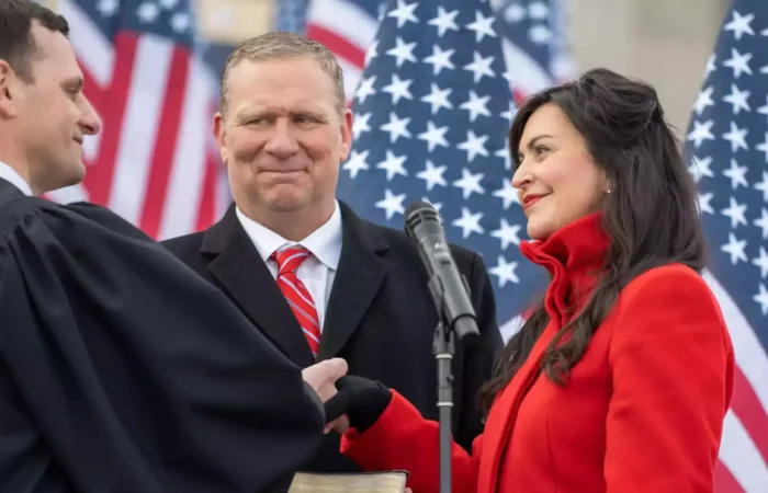 Secretary of State Christi Jacobsen sworn in to 2nd term during inauguration ceremony 5 At an inauguration ceremony, Christi Jacobsen, wearing a red coat, takes an oath with her hand on a Bible, smiling at the judge in a black robe, while a man in a suit and red tie stands beside her. American flags are displayed in the background.