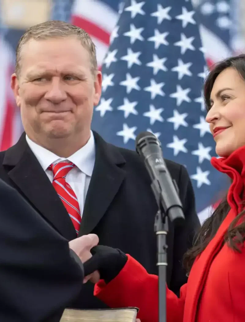 At an inauguration ceremony, Christi Jacobsen, wearing a red coat, takes an oath with her hand on a Bible, smiling at the judge in a black robe, while a man in a suit and red tie stands beside her. American flags are displayed in the background.