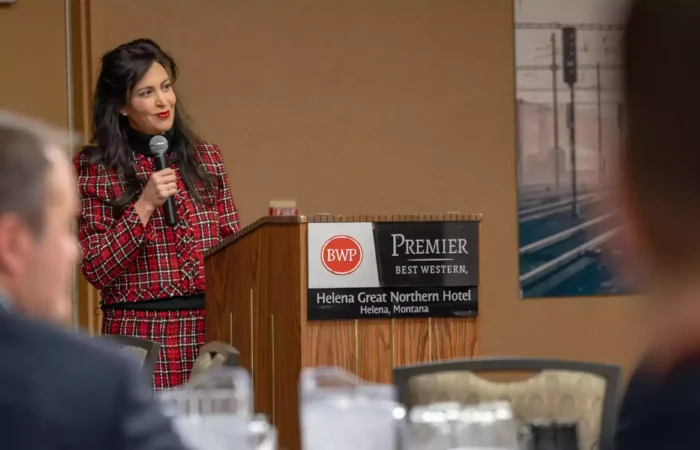 Secretary Christi Jacobsen: 'Montana's robust business growth continues in February' 1 A woman in a red plaid suit speaks at a podium with a microphone at the Helena Great Northern Hotel in Montana. The podium displays Best Western and hotel signage. People sit at tables in the foreground.