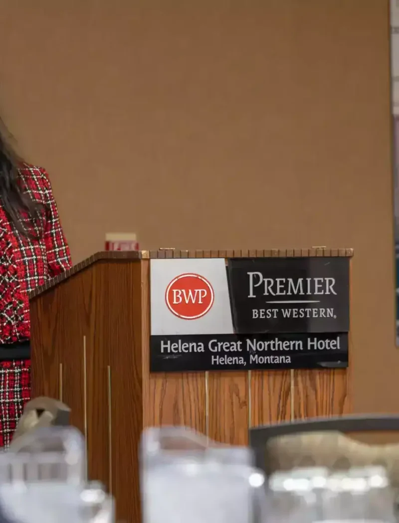 A woman in a red plaid suit speaks at a podium with a microphone at the Helena Great Northern Hotel in Montana. The podium displays Best Western and hotel signage. People sit at tables in the foreground.